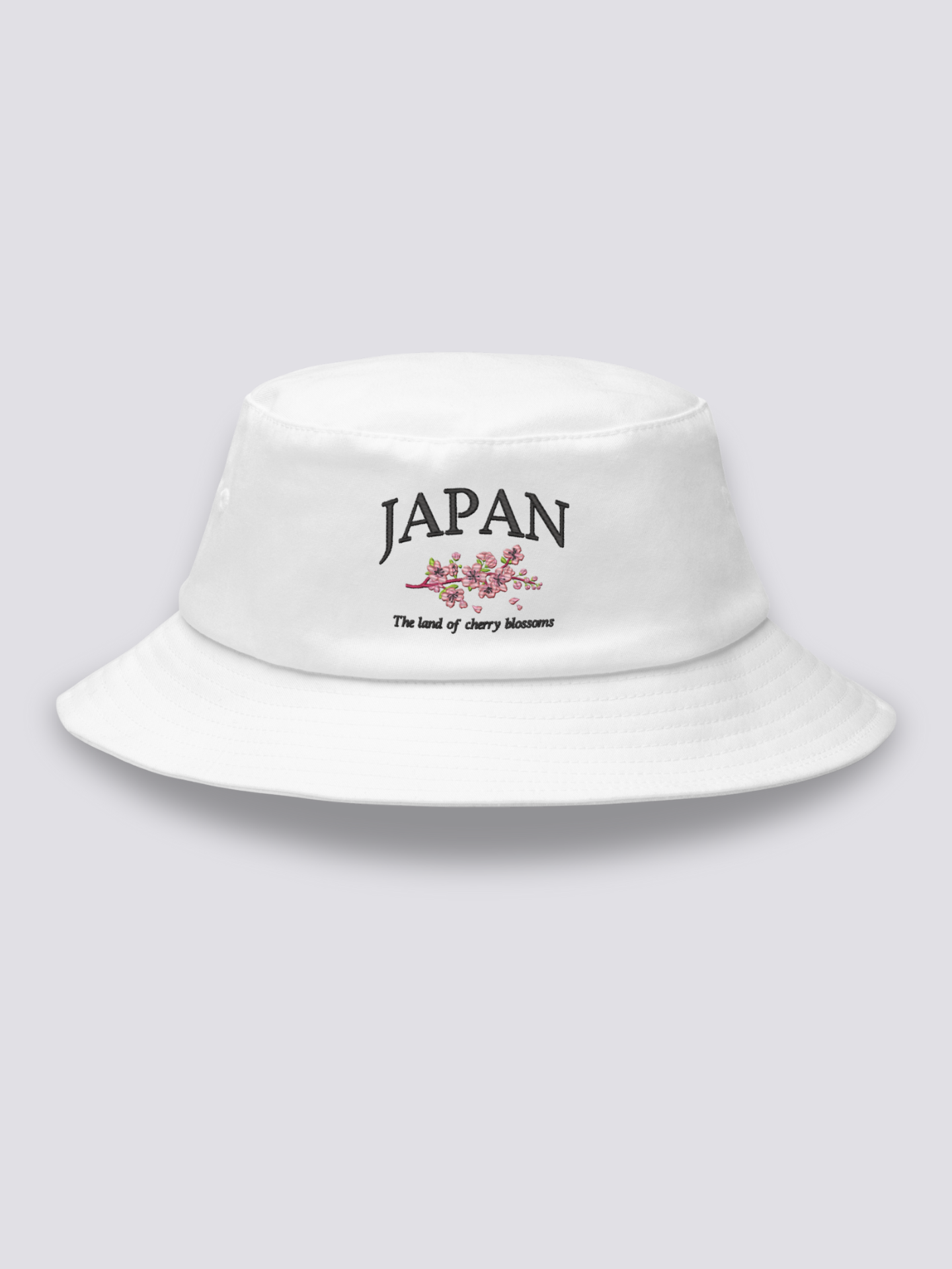 White bucket hat with JAPAN embroidered in black serif capitals crossed by a pink cherry branch above the line The land of cherry blossoms