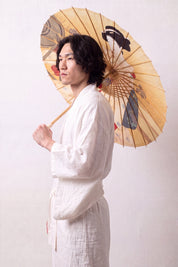 Traditional Japanese paper umbrella with a woman in a pale kimono with red accents on a glowing ochre-gold ground, held open overhead by a model in a white kimono