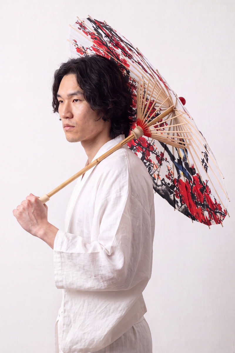 Traditional Japanese paper umbrella with an ink-brushed red plum branch and calligraphy on a white ground, held open by a model in a white kimono