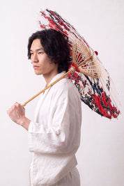 Traditional Japanese paper umbrella with an ink-brushed red plum branch and calligraphy on a white ground, held open by a model in a white kimono