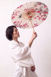 Traditional Japanese paper umbrella with a ring of pink magnolias and black swallows on a warm ivory canopy, held open overhead by a model in a white kimono
