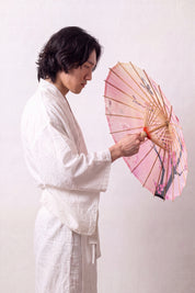 Traditional Japanese paper umbrella with fuchsia spring blossom and small sparrows on a peach-and-pink gradient, held open overhead by a model in a white kimono