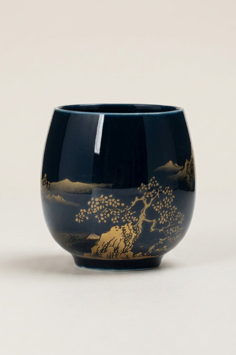 Handleless Japanese yunomi tea cup in dark navy-black glaze, decorated with a gold lakeside landscape of pine trees and mountains