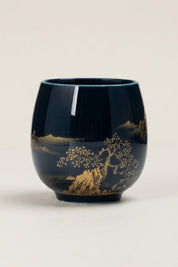 Handleless Japanese yunomi tea cup in dark navy-black glaze, decorated with a gold lakeside landscape of pine trees and mountains