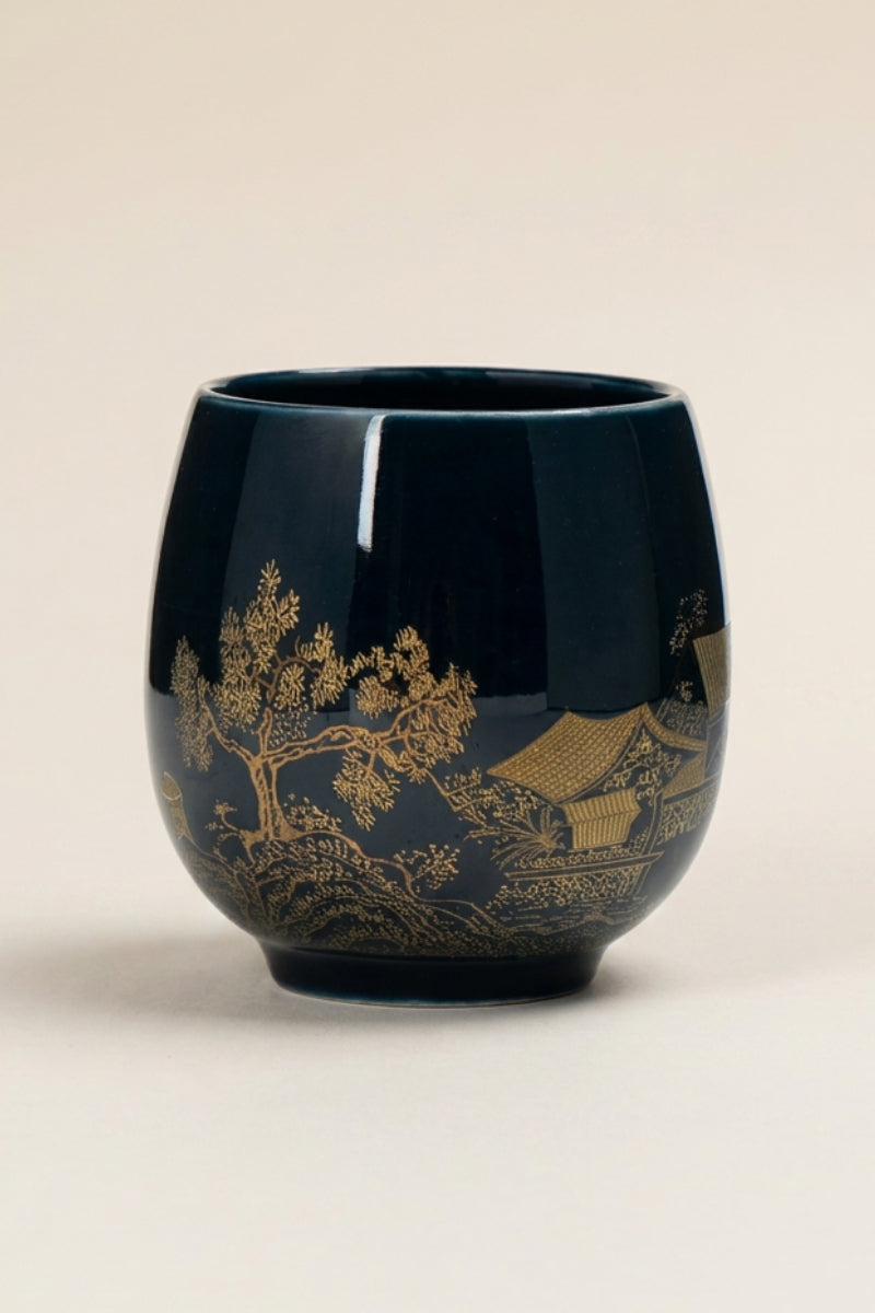 Handleless Japanese yunomi tea cup in dark navy-black glaze, decorated with a gold temple pavilion among pine trees