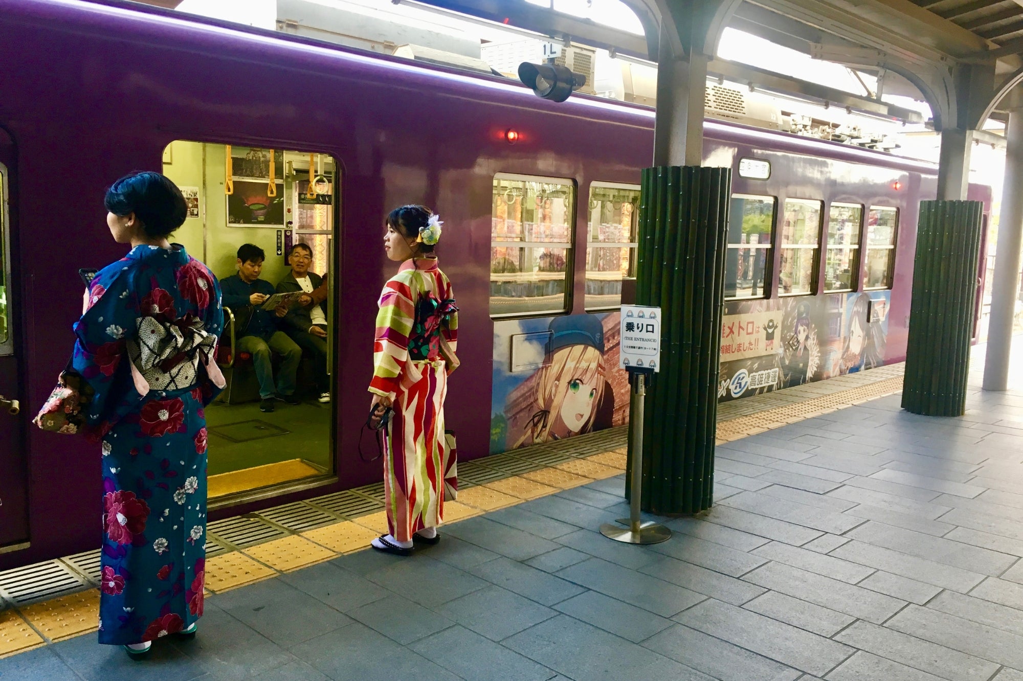 Two women in traditional kimonos waiting for an anime-themed train in Kyoto, illustrating the fusion of heritage and cosplay culture in the Japan Clothing Lookbook.