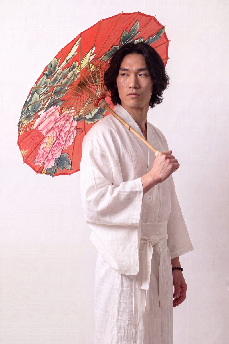 Traditional Japanese paper umbrella with large blush-pink peonies and green leaves on a bright red canopy, held open overhead by a model in a white kimono