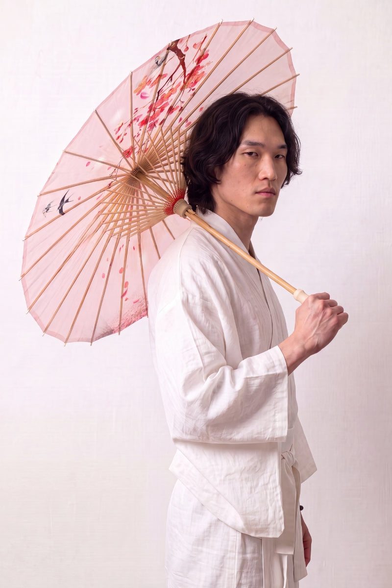Traditional Japanese paper umbrella with cherry blossom and two darting swallows on a blush-rose canopy, held open overhead by a model in a white kimono