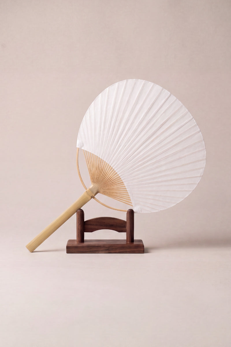 Flat white uchiwa fan with a rounded paper leaf on radiating bamboo ribs and a straight bamboo handle, shown on a wooden stand